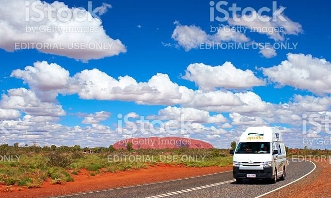 A white campervan on a sealed outback road with a red rock formation in the distance
